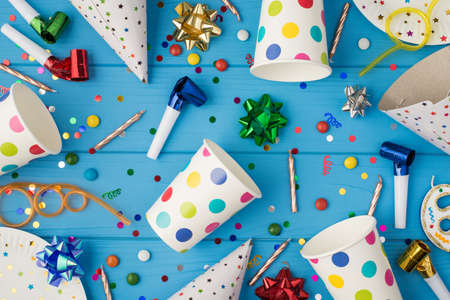 Top view photo of birthday party composition spiral cocktail tubes multicolored ribbon stars striped candles pipes hats confetti polka dots paper cups and plates isolated blue wooden table backgroundの写真素材