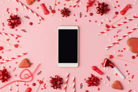 Top view photo of birthday composition with red ribbon stars serpentine confetti bright decorations and smartphone with copyspace in the middle on pastel pink backgroundの写真素材