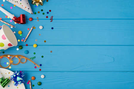Top view photo of birthday party composition spiral tube ribbon stars candles pipes hat confetti polka dots paper cup plate on the left on isolated blue wooden table background copyspace on the rightの写真素材