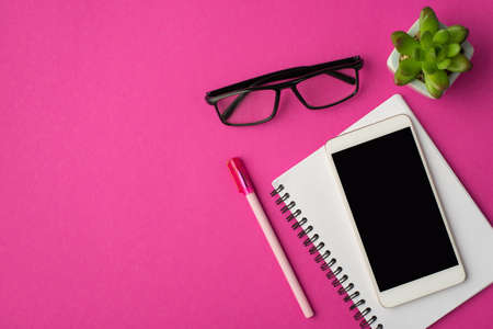 Top view photo of workplace with smartphone on notebook plant pen and glasses on isolated pink background with blank spaceの写真素材