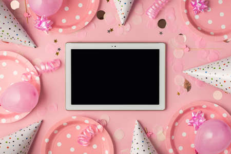 Top view photo of birthday composition with pink ribbon stars serpentine hats paper plates balloons confetti and tablet computer in the middle with copyspace on pastel pink backgroundの写真素材