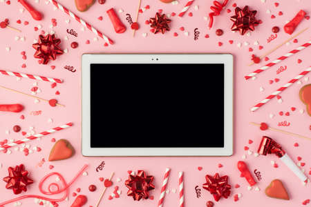 Top view photo of birthday composition with red ribbon stars serpentine confetti bright decorations and tablet computer in the middle with copyspace on pastel pink backgroundの写真素材