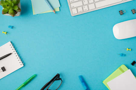 Top view photo of desktop with keyboard pens clips organizes pencil on stickers pen and clip on notebook glasses mouse and plant on isolated blue background with copyspaceの写真素材