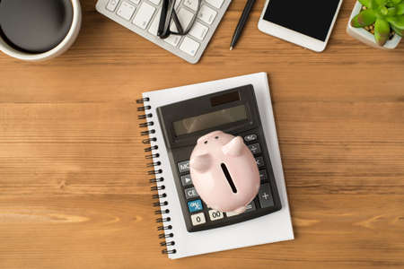 Overhead photo of notebook calculator piggybank glasses plant pen phone keyboard and cup of coffee isolated on the wooden backdropの写真素材