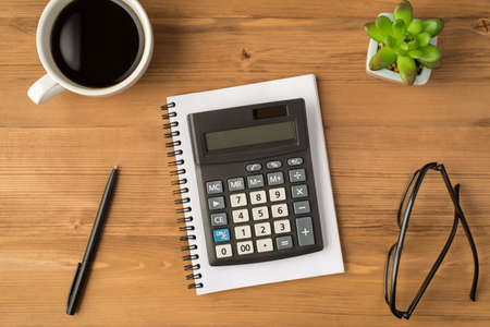 Overhead photo of notebook calculator glasses plant pen and cup of coffee isolated on the wooden backdropの写真素材
