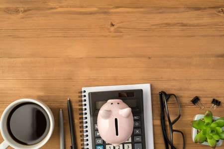 Overhead photo of notebook calculator glasses two pens piggybank plant paperclips and cup of coffee isolated on the wooden backdropの写真素材