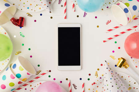 Above photo of cups plate confetti birthday hat party tubules whistle balloons and empty phone isolated on the white backgroundの写真素材