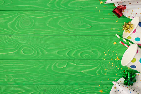 Top view photo of birthday party composition on the right ribbon stars pipe straws hats sequins polka dots paper cups and plates on isolated green wooden table background with copyspace on the leftの写真素材