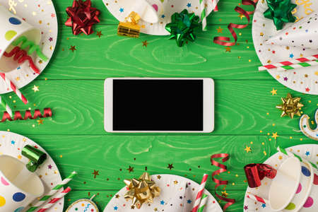 Top view photo of birthday party composition horizontally lying smartphone ribbon stars spiral candles pipes straws hat sequins paper cups and plates on isolated green wooden table background