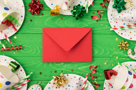 Top view photo of birthday party composition closed red envelope in the middle ribbon stars candles pipes straws hat sequins polka dots paper cups and plates on isolated green wooden table backgroundの写真素材