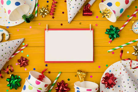 Above photo of cups plate confetti birthday hat party tubules whistle bow invitation card paper isolated on the yellow wooden backgroundの写真素材