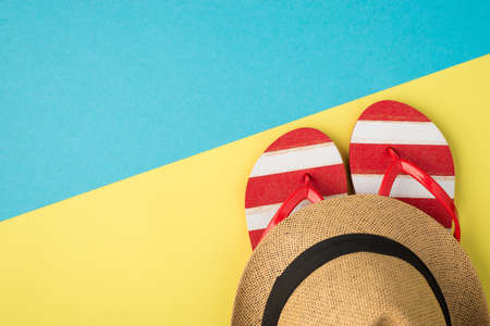 Top view photo of hat on striped red and white flip-flops on bicolor yellow and blue background with...