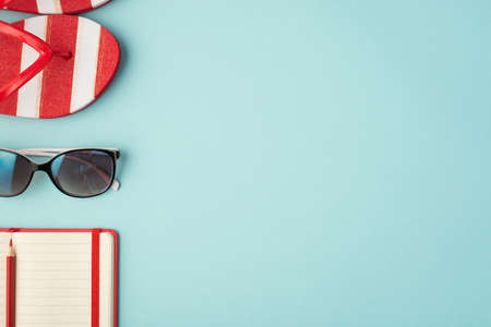 Top view photo of striped flip-flops sunglasses and red planner with pencil on isolated pastel blue background with copyspaceの写真素材