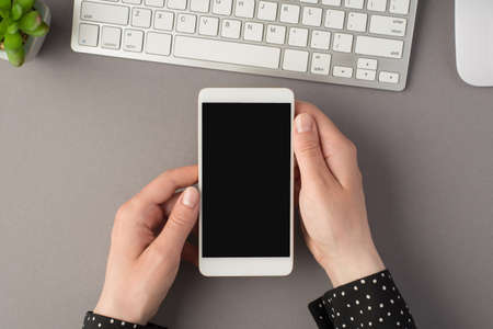 First person top view photo of white keyboard mouse flowerpot and woman's hands holding smartphone...