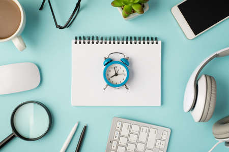 Top view photo of alarm clock on horizontal planner cup white keyboard mouse magnifier pens headphones smartphone plant and glasses on isolated pastel blue background with blank spaceの写真素材