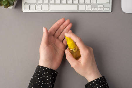 First person top view photo of female hands using sanitizer spray yellow bottle white keyboard mouse and flowerpot on isolated gray backgroundの写真素材