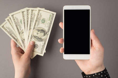 First person top view photo of woman's hands holding smartphone and fan of money hundred dollar banknotes on isolated gray background with copyspaceの写真素材