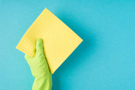 Top view photo of hand in green rubber glove holding yellow viscose napkin on isolated pastel blue...
