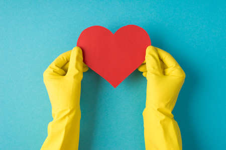 Top view photo of hands in yellow rubber gloves holding red paper heart on isolated pastel blue...