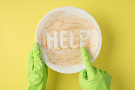 Top view photo of hands in green rubber gloves indicating with forefinger on dirty plate with inscription help on isolated yellow backgroundの写真素材
