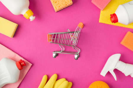 Top view photo of shopping cart model in the middle scouring pads rags rubber gloves and white detergent bottles on isolated pink backgroundの写真素材