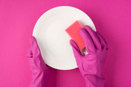 Top view photo of hands in pink gloves holding white clean dish and pink sponge on isolated pink backgroundの写真素材