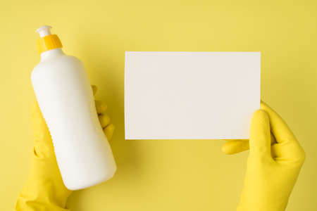 Top view photo of hands in yellow rubber gloves holding white detergent bottle without label and...