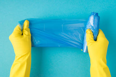 Top view photo of hands in yellow rubber gloves unfolding blue garbage bags on isolated pastel blue...