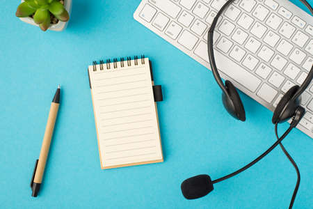 Top view photo of black headset with microphone on white keyboard flowerpot pen and reminder on isolated pastel blue background with blank spaceの写真素材