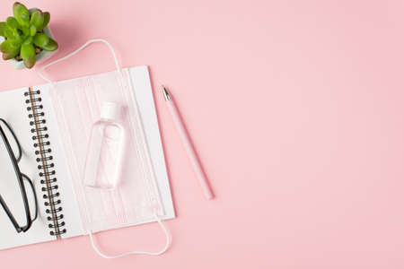 Top view photo of pink medical facemask sanitizer glasses on open notebook pen and flowerpotの写真素材