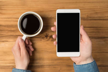 Overhead photo of smartphone in hands and cup of coffee isolated on the wooden backdropの写真素材
