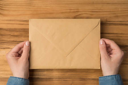 First person top view photo of hands holding closed craft paper envelope on isolated wooden table background with copyspaceの写真素材