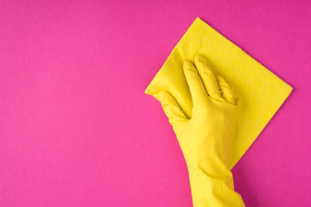 Top view photo of hand in yellow glove using yellow viscose rag on isolated pink background with...