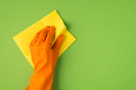 Above photo of hands in orange gloves and yellow cleaning rag isolated on the green background