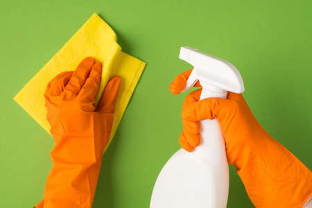 Overhead photo of hands in orange gloves holding cleaning spray and cleaning rag isolated on the green backgroundの写真素材
