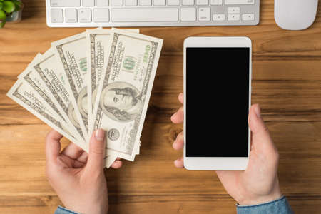 First person top view photo of female hands holding fan of money hundred dollars banknotes and smartphone flowerpot white keyboard and mouse on isolated wooden table background with copyspaceの写真素材