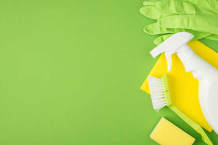 Top view photo of white spray detergent bottle sponge brush yellow napkin and green rubber gloves on isolated green background with copyspace on the leftの写真素材