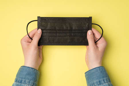 First person top view photo of woman's hand holding black medical facemask on isolated yellow background with blank spaceの写真素材