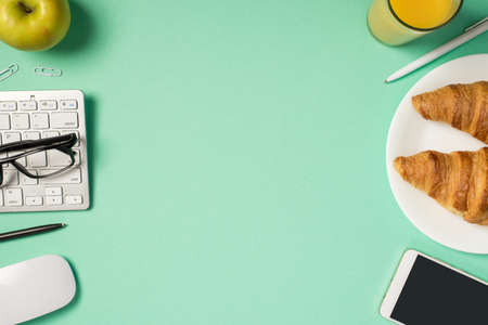 Top view photo of workplace keyboard glasses mouse pens clips smartphone apple glass of beverage and plate with two croissants on isolated turquoise background with blank space in the middleの写真素材