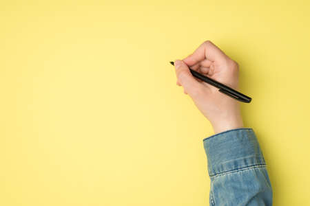 First person top view photo of woman's hand holding black pen on isolated yellow background with blank spaceの写真素材