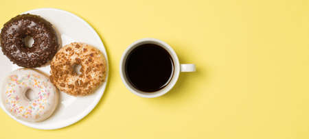 Top view photo of cup of coffee and plate with three different donuts on isolated light yellow...