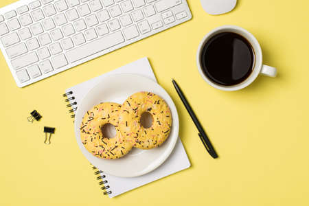 Top view photo of workplace white keyboard mouse plate with two yellow glazed donuts on spiral notebook cup of coffee binder clips and pen on isolated light yellow backgroundの写真素材