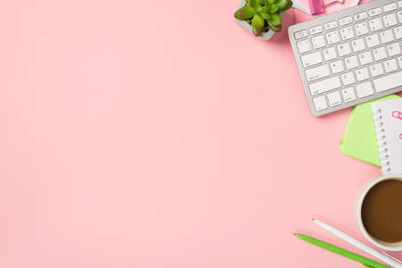 Above photo of keyboard cup of coffee notepad pens paper clips and green plant isolated on the pink...