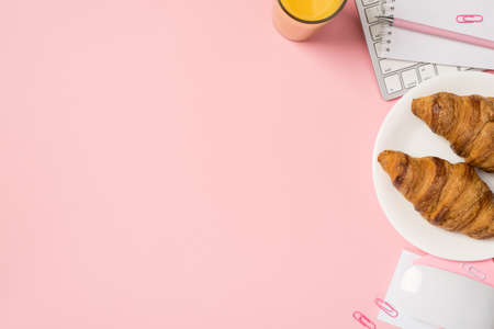 Overhead photo of keyboard computer mouse glass of orange juice notepad with pen paper clips and plate with two croissants isolated on the pink backdrop with empty spaceの写真素材
