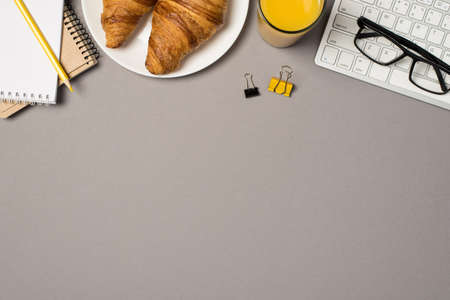 Top view photo of workplace keyboard glasses binder clips notepads pencil glass of juice and plate with fresh croissants on isolated gray background with copyspaceの写真素材