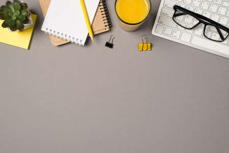 Top view photo of workplace glasses on keyboard glass of orange juice binder clips yellow pencil on planners and flowerpot on sticker note paper on isolated gray background with empty spaceの写真素材