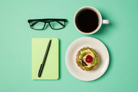 Overhead photo of cup of coffee notepad with pen glasses and plate with fruit cake isolated on the...