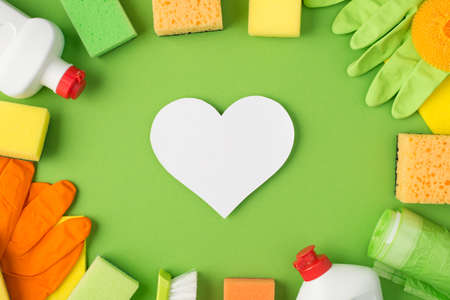 Top view photo of white paper heart in the middle detergent bottles rubber gloves rags scouring pads brush and garbage bags on isolated green background with copyspaceの写真素材