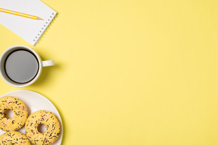 Top view photo of cup of drink plate with yellow glazed donuts and yellow pencil on small notebook...