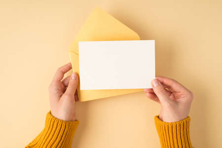 First person top view photo of woman's hands in yellow pullover holding open pastel yellow envelope and white card on isolated light orange background with blank spaceの写真素材